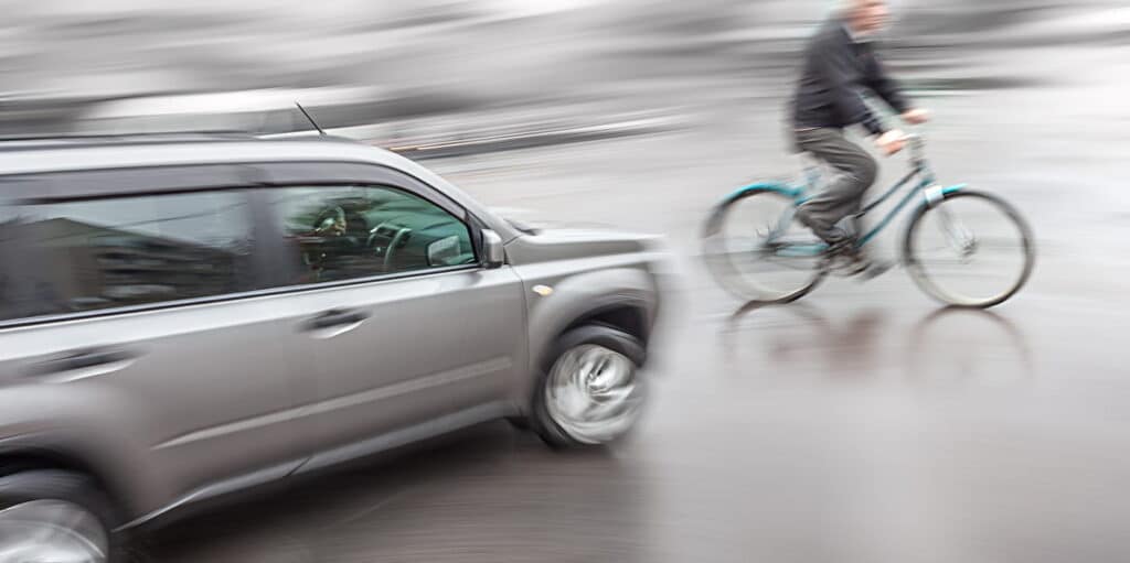 A blurred image of a vehicle almost about to hit a person on a bicycle.