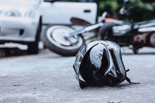 A helmet on the pavement with a crashed motorcycle and vehicle in the background.