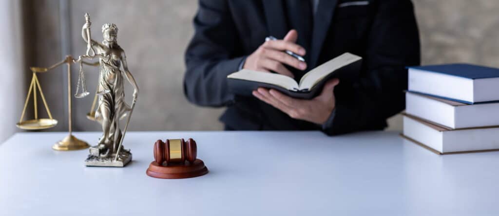 A lawyer writing in a book at his desk. In front of him are more books, a gavel, a Lady Justice statue, and the scales of justice.