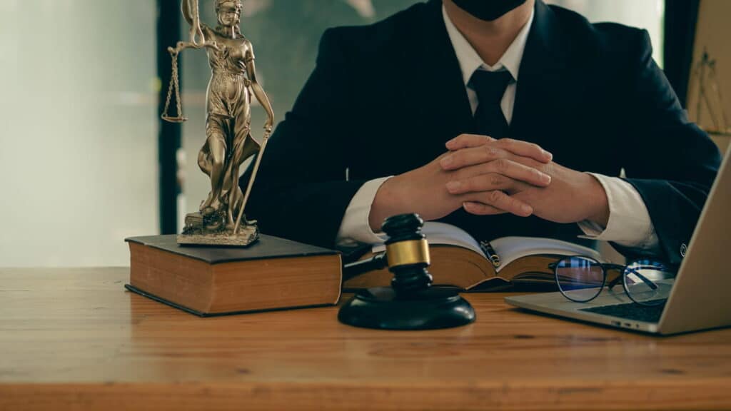 A personal injury attorney sits at his desk with his hands together. In front of him is an open laptop with a pair of glasses on them, an open book, a gavel, and a Lady Justice statue propped on a closed book.
