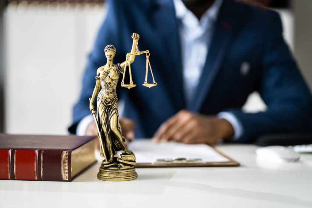 Focus is on a lady justice statue next to a book as a car accident lawyer works on paperwork in the background.