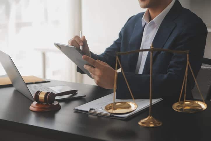 A medical malpractice lawyer working on a tablet at his desk. In front of him is paperwork, an open laptop, a gavel, and the scales of justice.