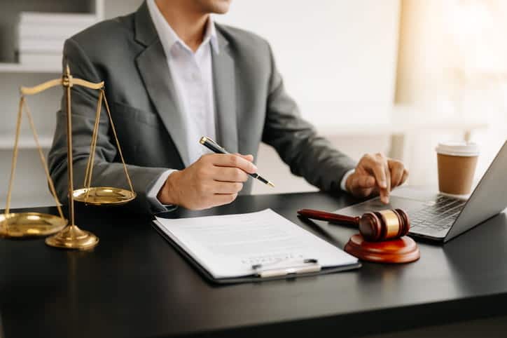 A personal injury lawyer working on paperwork at his desk. Next to him is a cup of coffee, an open laptop, a gavel and the scales of justice. 