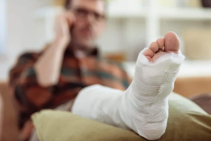 A man out of focus on his cellphone while he rests his broken leg on a pillow after suffering from a personal injury.