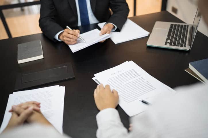 A personal injury lawyer reviewing paperwork with his two clients. On his desk is more paperwork, a book, and an open laptop. 