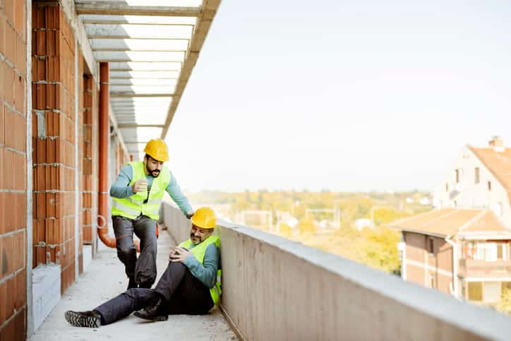 A construction worker is rushing over to another worker who is sitting on the ground, clutching his knee in pain after an accident. 