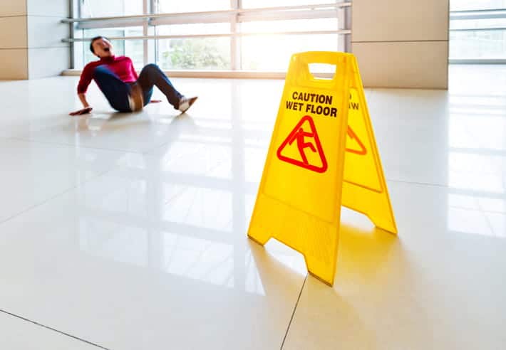 A man in pain on the ground after slipping and falling near a yellow caution wet floor sign.