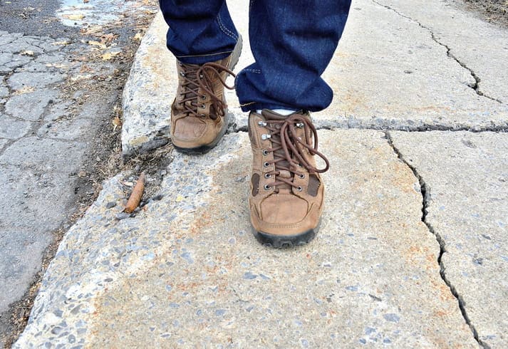 Focus is on a person's feet as they walk over a damaged and cracked sidewalk.