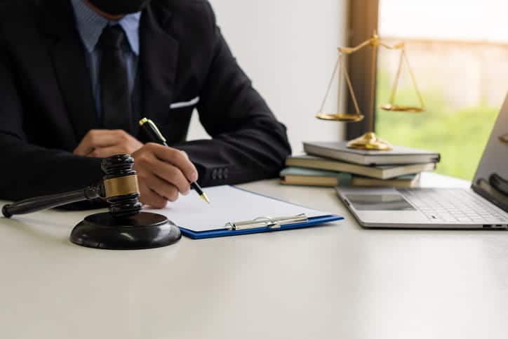 A car accident attorney works on paperwork at his desk. Next to him is a stack of books, the scales of justice, an open laptop, and a gavel.