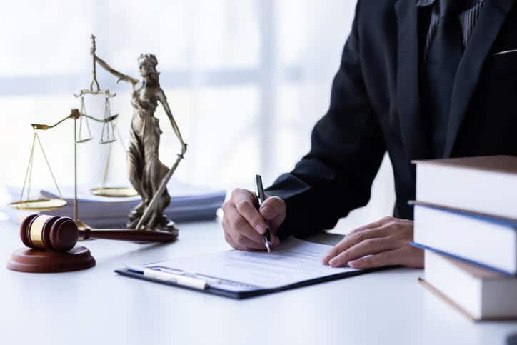 A car accident lawyer working on paperwork at his desk with a stack of books, a Lady Justice statue, the scales of justice and a gavel next to him. 