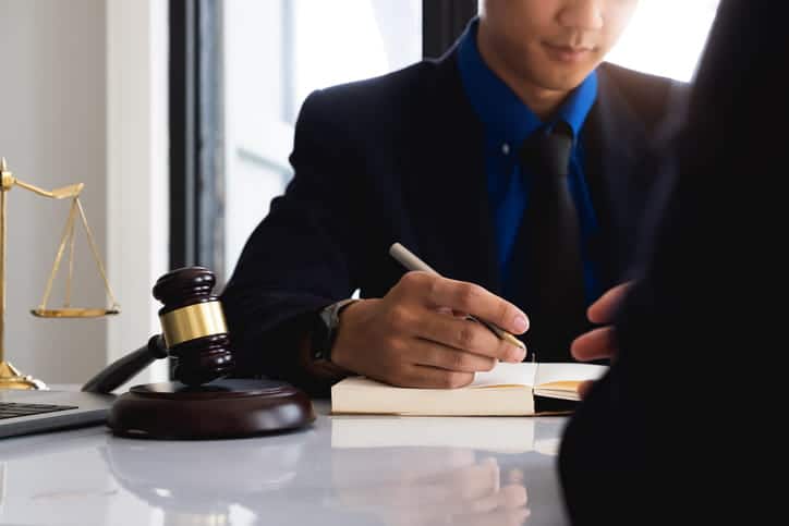 A personal injury lawyer writing in a notebook at his desk. Next to him is the scales of justice and a gavel. 