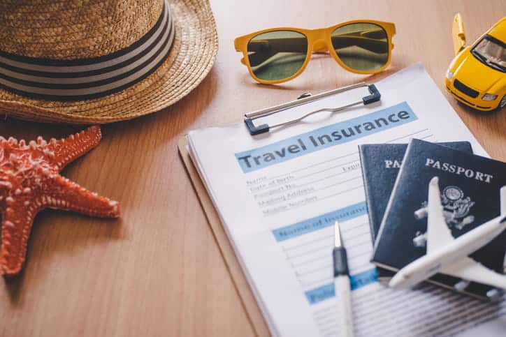 A clipboard with papers labeled "travel insurance. " surrounding it is a starfish, a hat, sunglasses, a toy plane and car, with two passports and a pen.